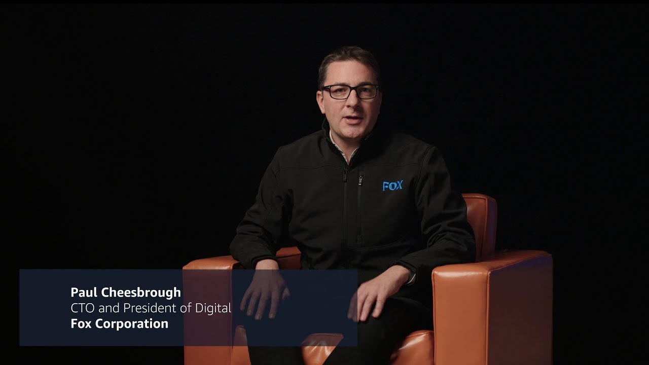 YouTube video thumbnail featuring a person seated in an orange chair, wearing a FOX-branded jacket, with a caption overlay reading 'Paul Cheesbrough, CTO and President of Digital, Fox Corporation.'