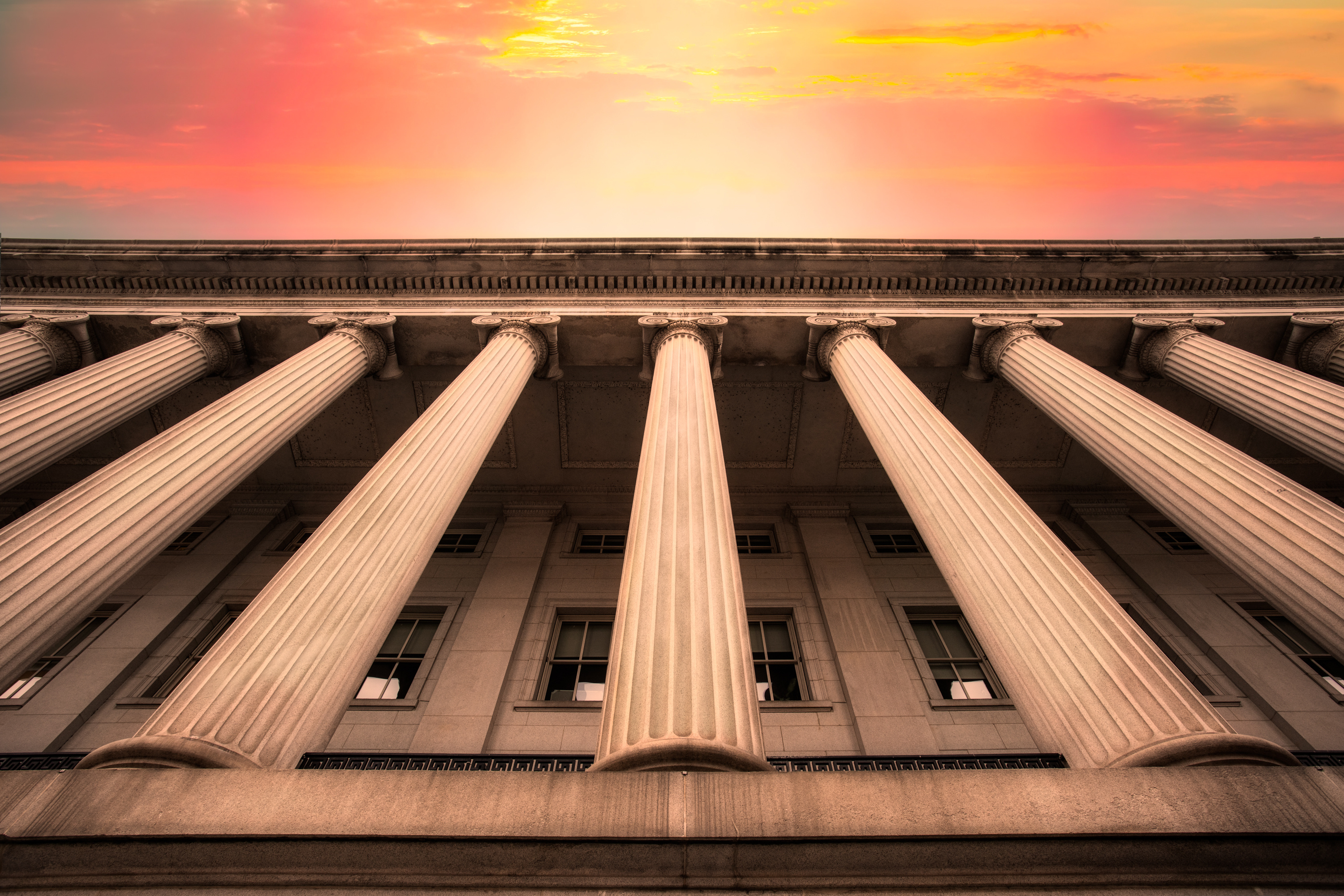 A low-angle view of neoclassical building columns in front of a facade, with a dramatic sunset sky in the background.