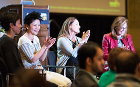 A woman in a red blazer speaks on a panel while others applaud in a conference setting.