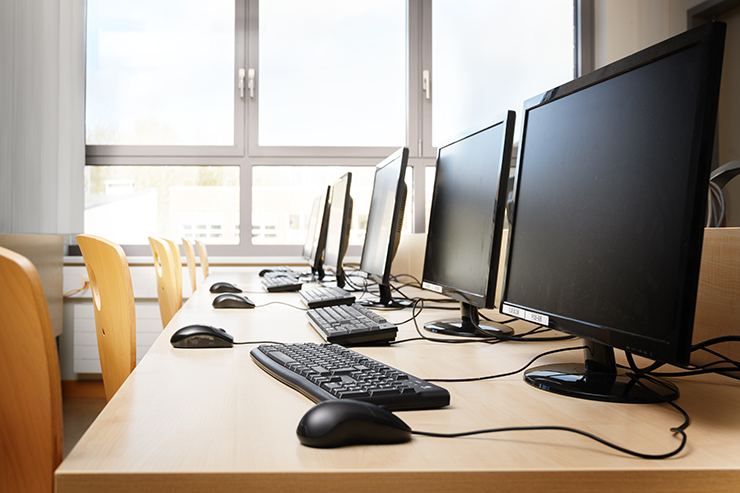 Row of desktop computers with keyboards and mice on a wooden table in a bright classroom.