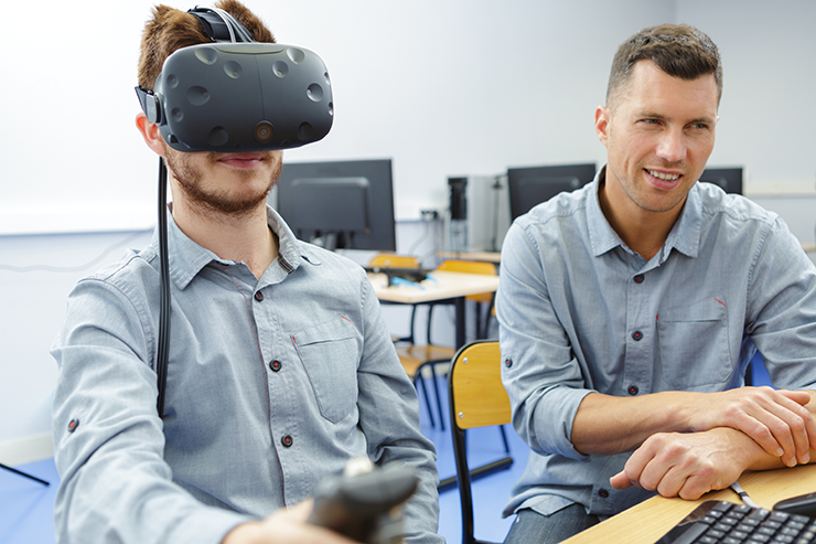 Two people in a classroom setting, one wearing a virtual reality headset and holding a controller, with computers visible in the background.
