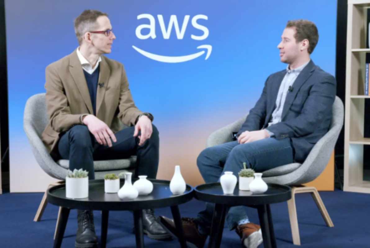Two people sit in modern chairs on a studio set with the AWS logo in the background, having a discussion. Small vases and plants are on tables between them. This image is used as the thumbnail for a video interview about AWS Nitro.