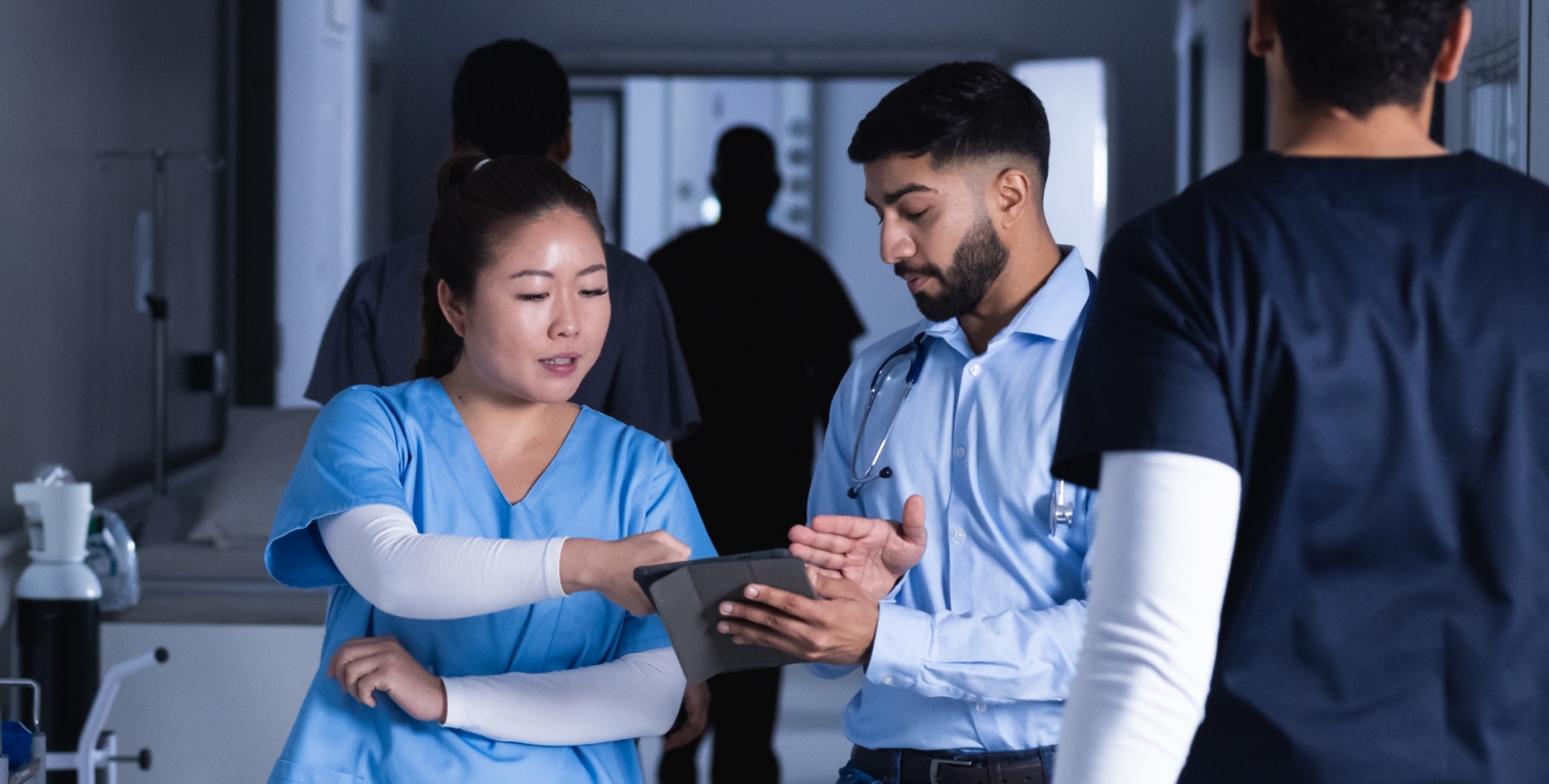 A group of healthcare professionals collaborate and discuss patient information using a tablet in a hospital hallway.
