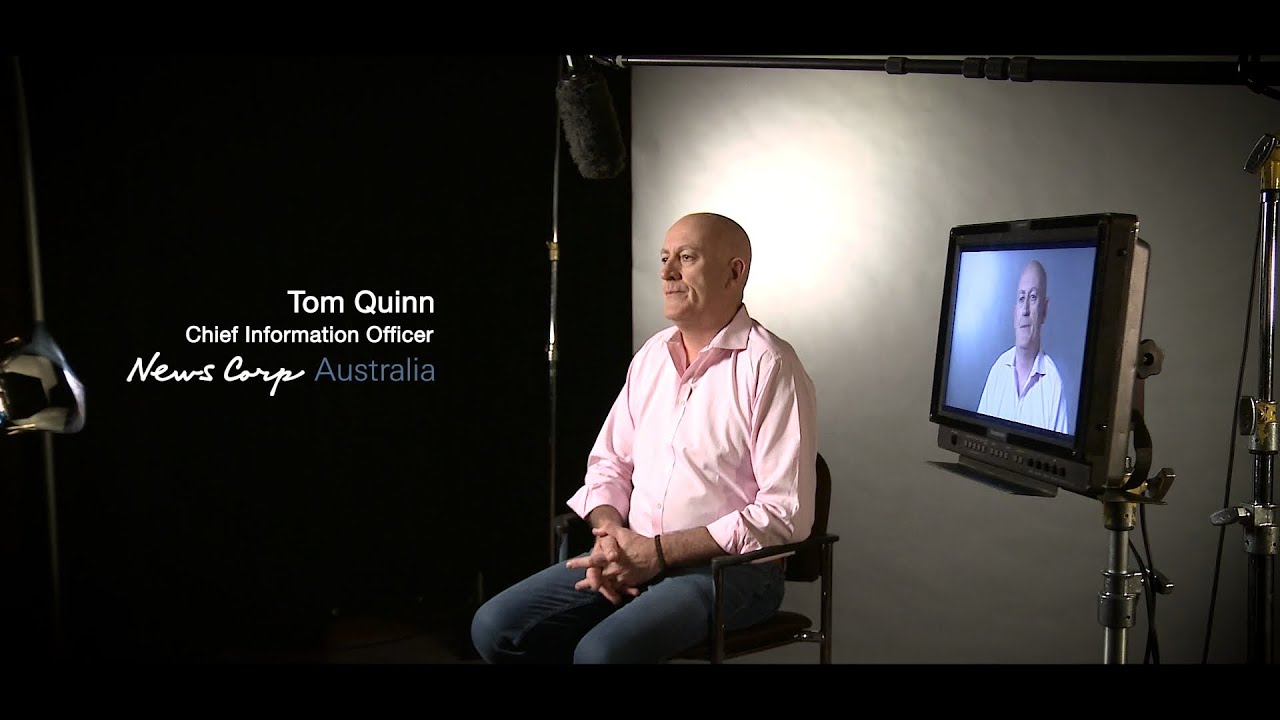 A man in a pink shirt, seated for an interview in a studio setting, with video production equipment visible and a monitor displaying his image. The text on screen indicates he is the Chief Information Officer of News Corp Australia.