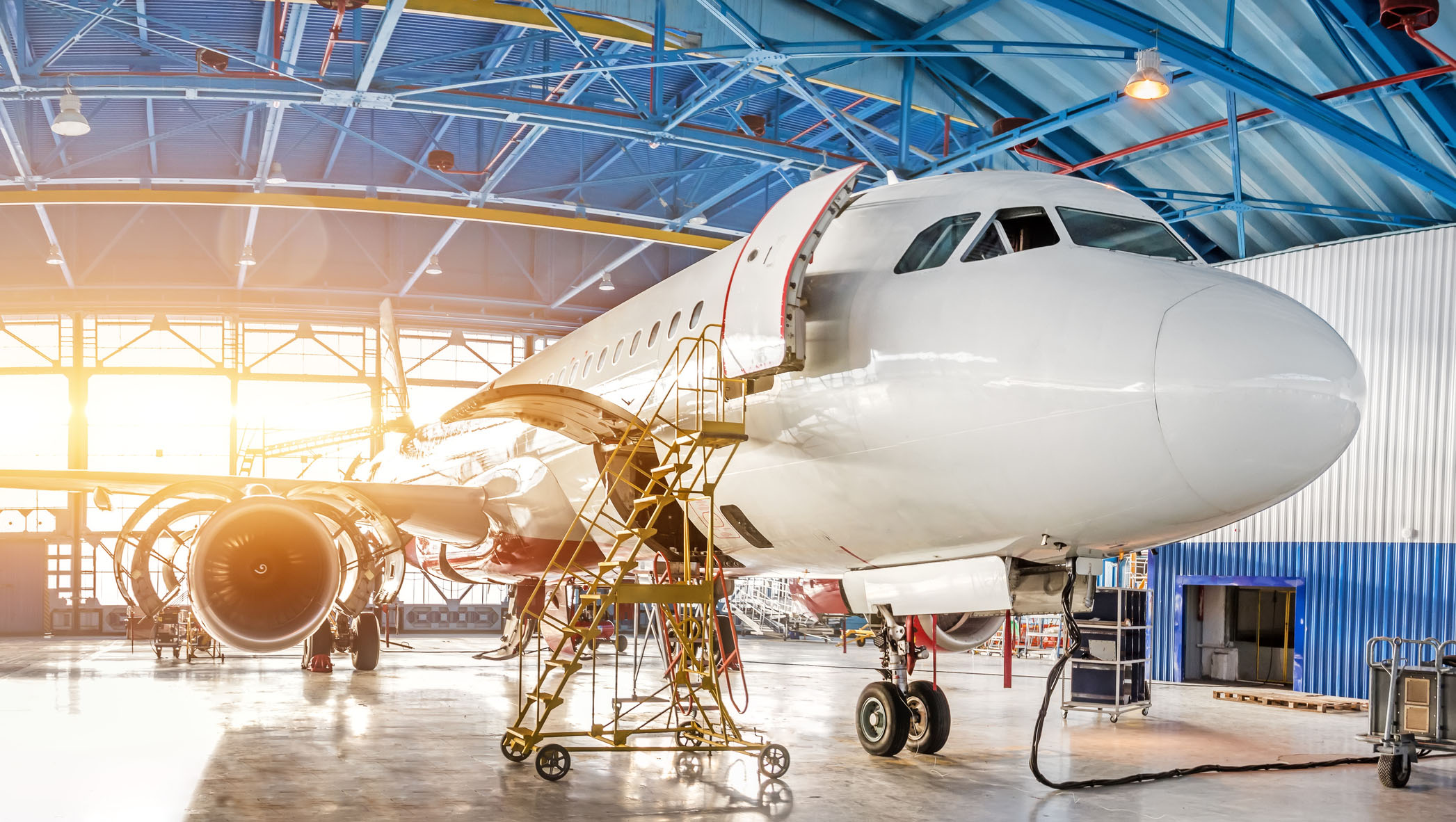 A commercial airplane undergoing maintenance inside a spacious hangar, with access stairs and equipment positioned around the aircraft. Sunlight shines through large windows, illuminating the scene and highlighting the engineering environment.