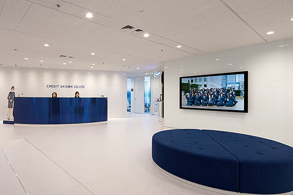 Reception area of Credit Saison office featuring a modern blue front desk, two receptionists, white interiors, a large circular blue seating area, and a wall-mounted screen displaying a group photo.