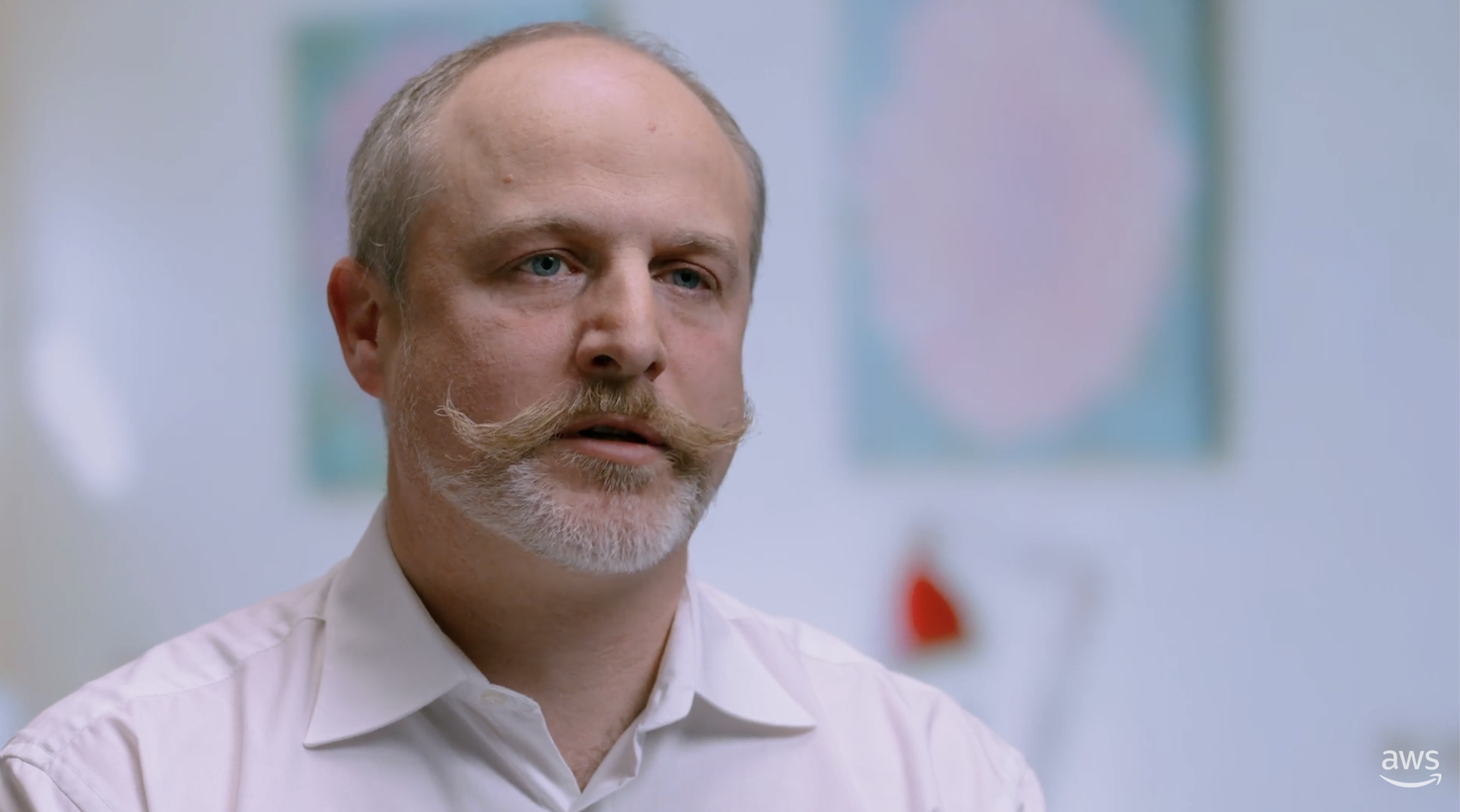 A business professional with a prominent mustache and beard, wearing a white shirt, is speaking in what appears to be an interview or testimonial setting. The background is softly blurred with pastel artwork and an office environment visible. AWS logo is present in the bottom right corner.