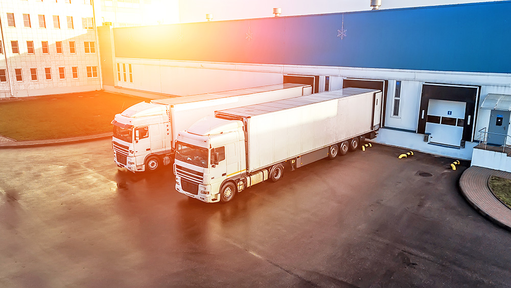 Two large white semi-trailer trucks parked at a distribution center loading dock on a sunny day, ready for cargo loading or unloading.