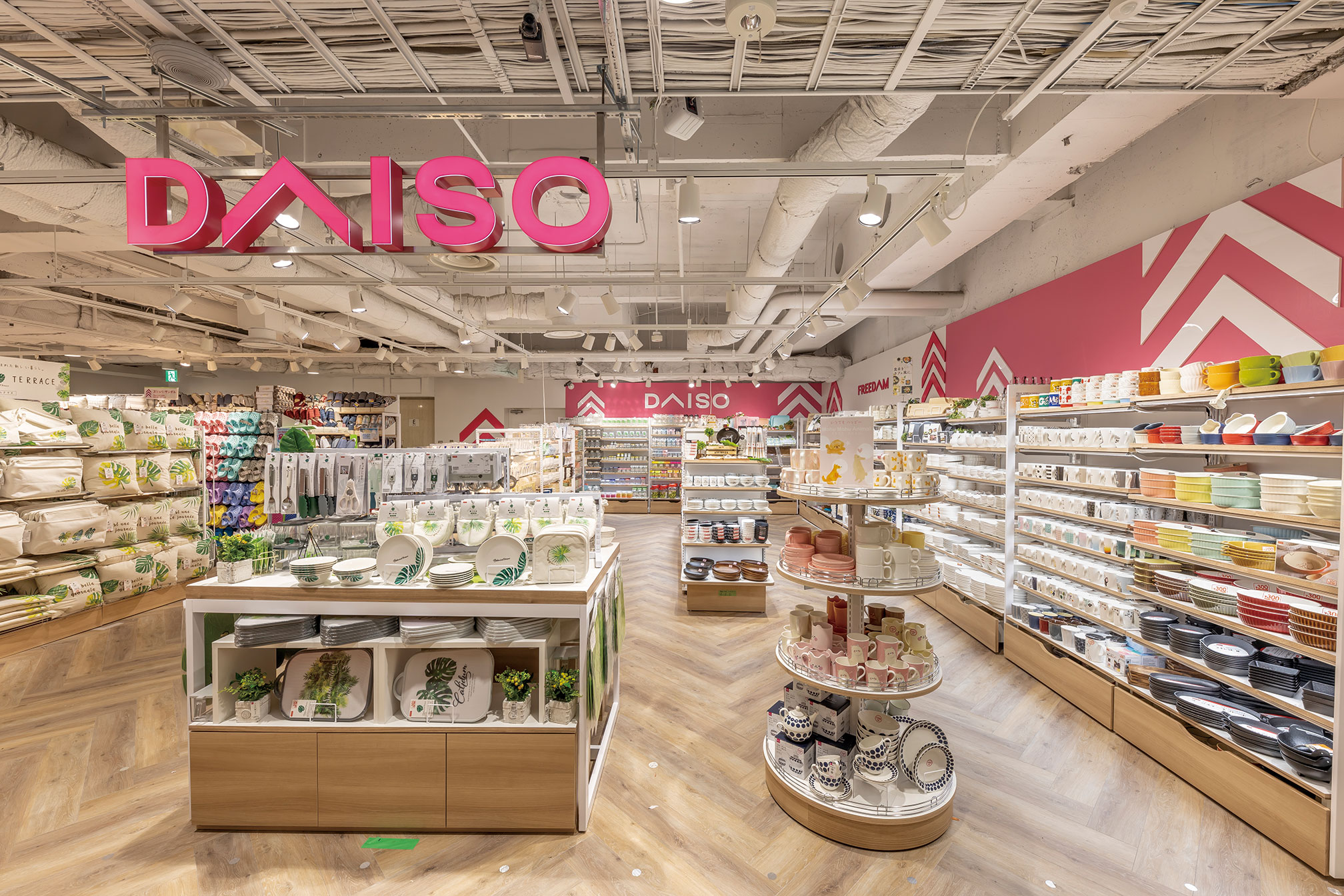 Wide-angle view of the inside of a DAISO retail store, featuring shelves filled with household items, kitchenware, dishes, and various colorful products neatly arranged under bright lighting and modern decor.