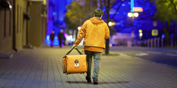 A delivery worker wearing an orange jacket walks down a city street at night carrying an insulated delivery bag. The urban setting is illuminated with vibrant blue and yellow lights, and the street appears calm and empty.