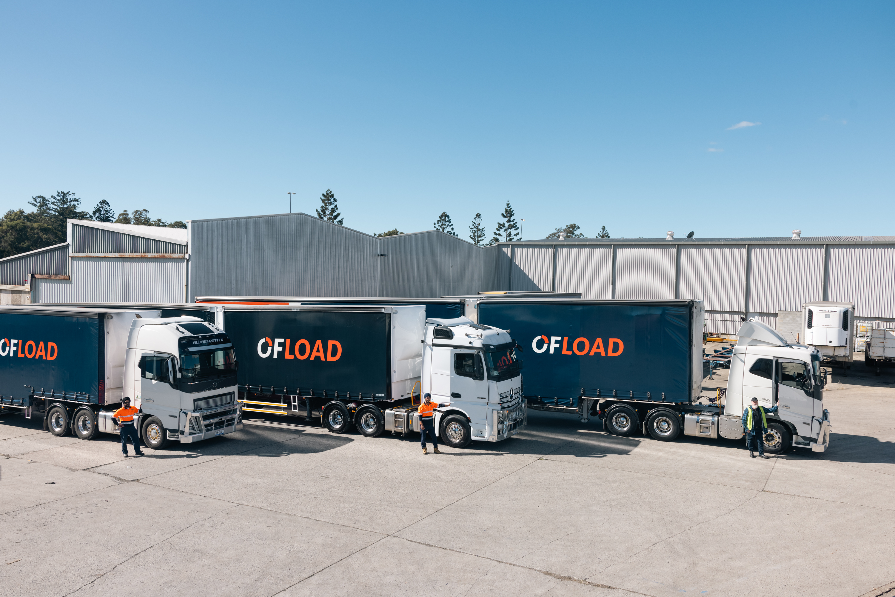 Three semi-trucks with "OFLOAD" branding parked in an industrial lot, with workers in high-visibility clothing standing beside them under a clear blue sky.