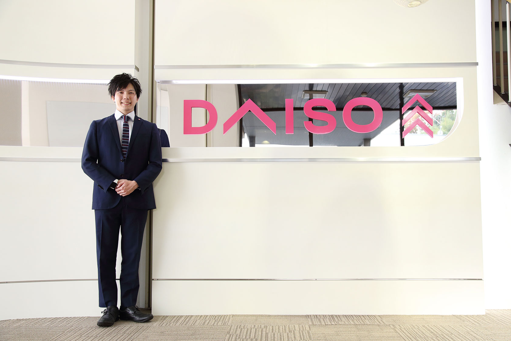 A person in a business suit stands next to the reception desk with the DAISO logo displayed in pink on the wall behind.