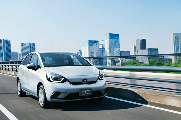 A white Honda Fit car driving on an urban highway with modern city buildings in the background under a clear blue sky.
