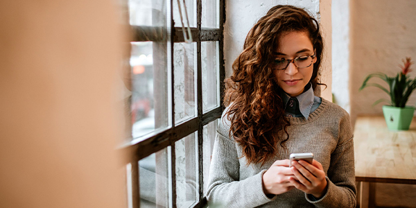 A woman with curly hair and glasses is sitting by a window in a cafe, wearing a sweater and looking at her smartphone.
