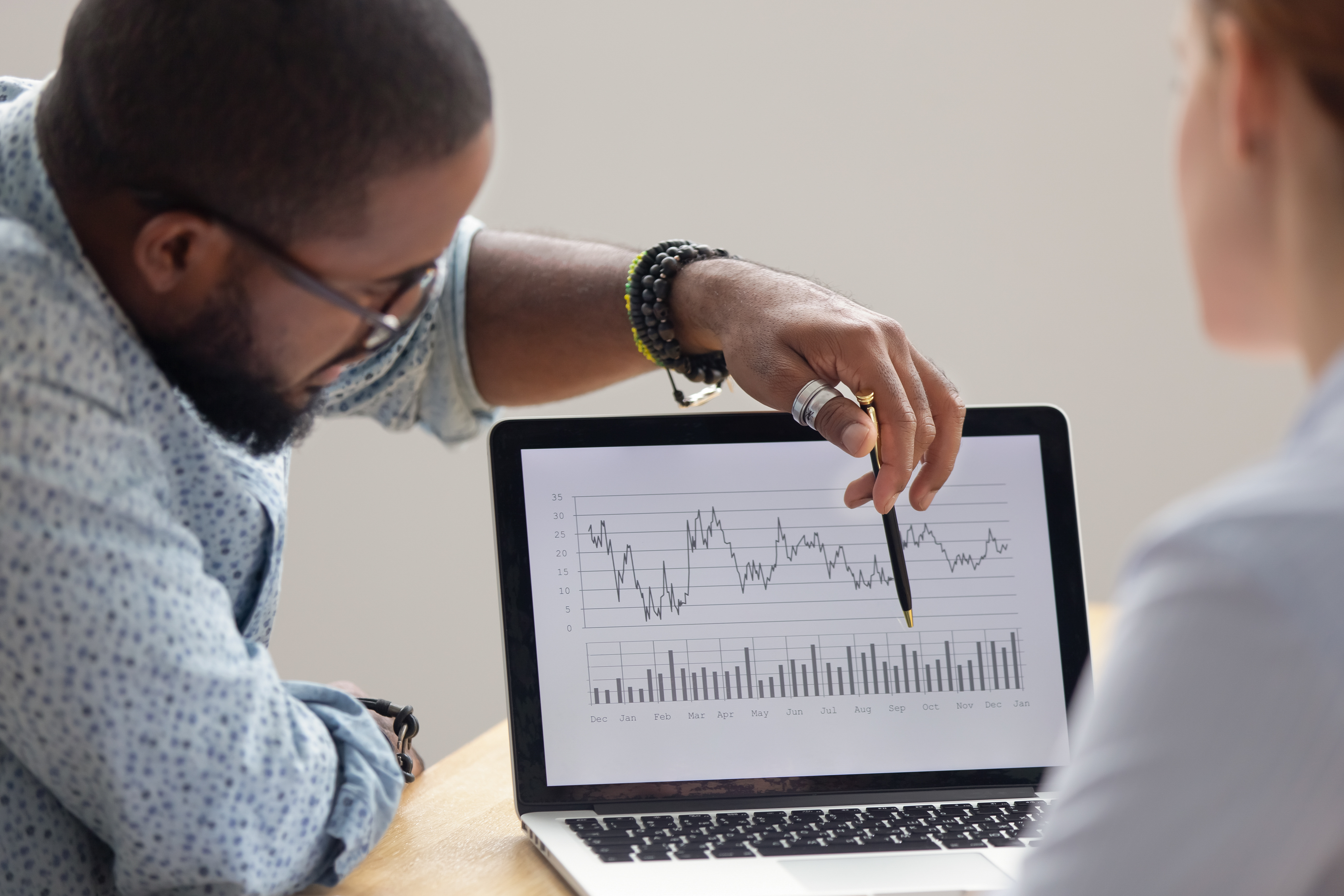 Focused african analyst showing client or colleague annual financial report analyzing business data on laptop screen using software for digital graphic statistic analysis, economic market graphs on computer, rear close up view