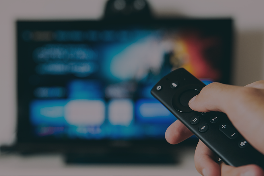 A hand holding a smart TV remote control in front of a television screen displaying the Prime Video interface.