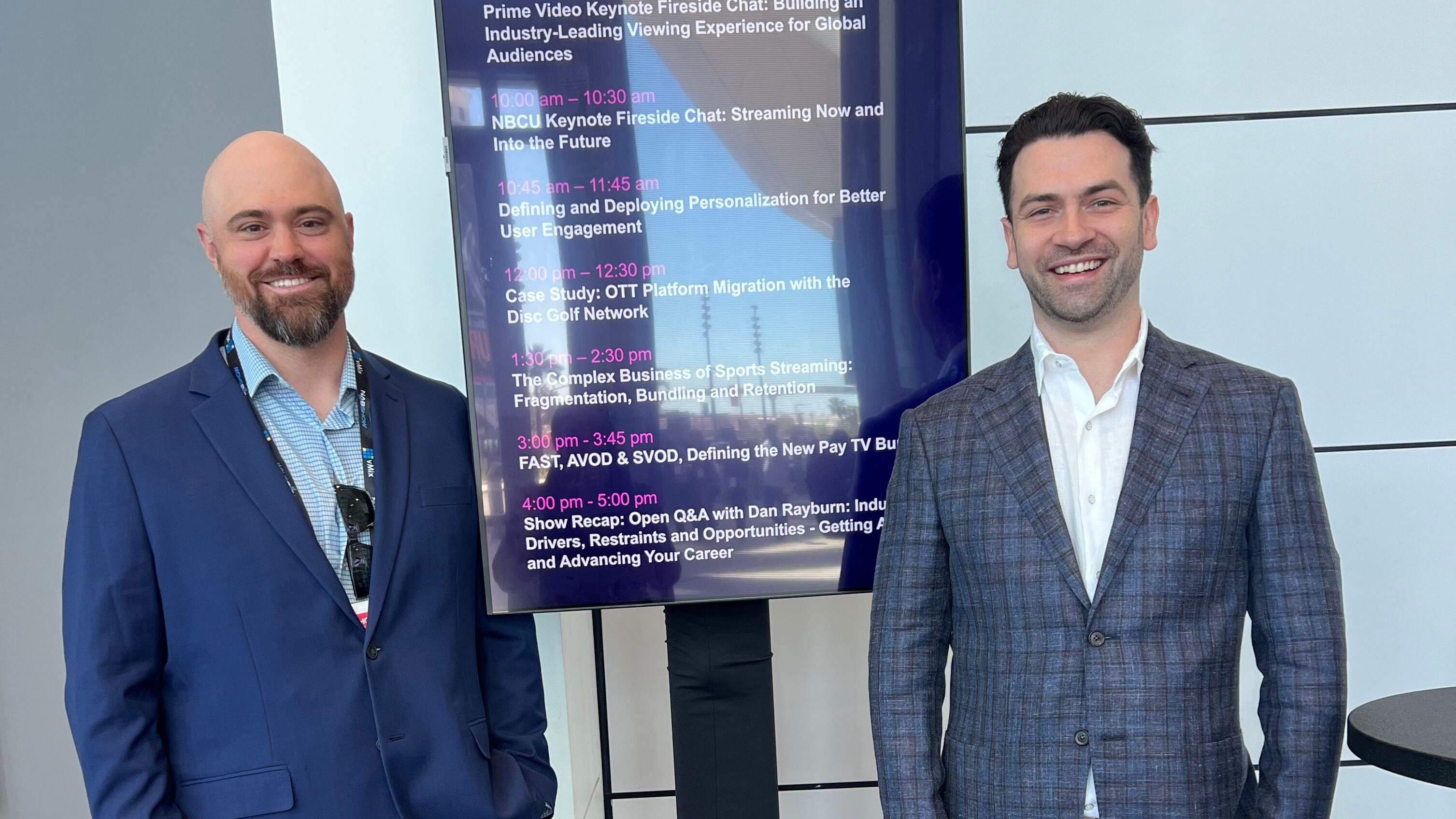 Two individuals in business attire standing in front of a digital schedule display listing conference sessions on streaming and media topics.