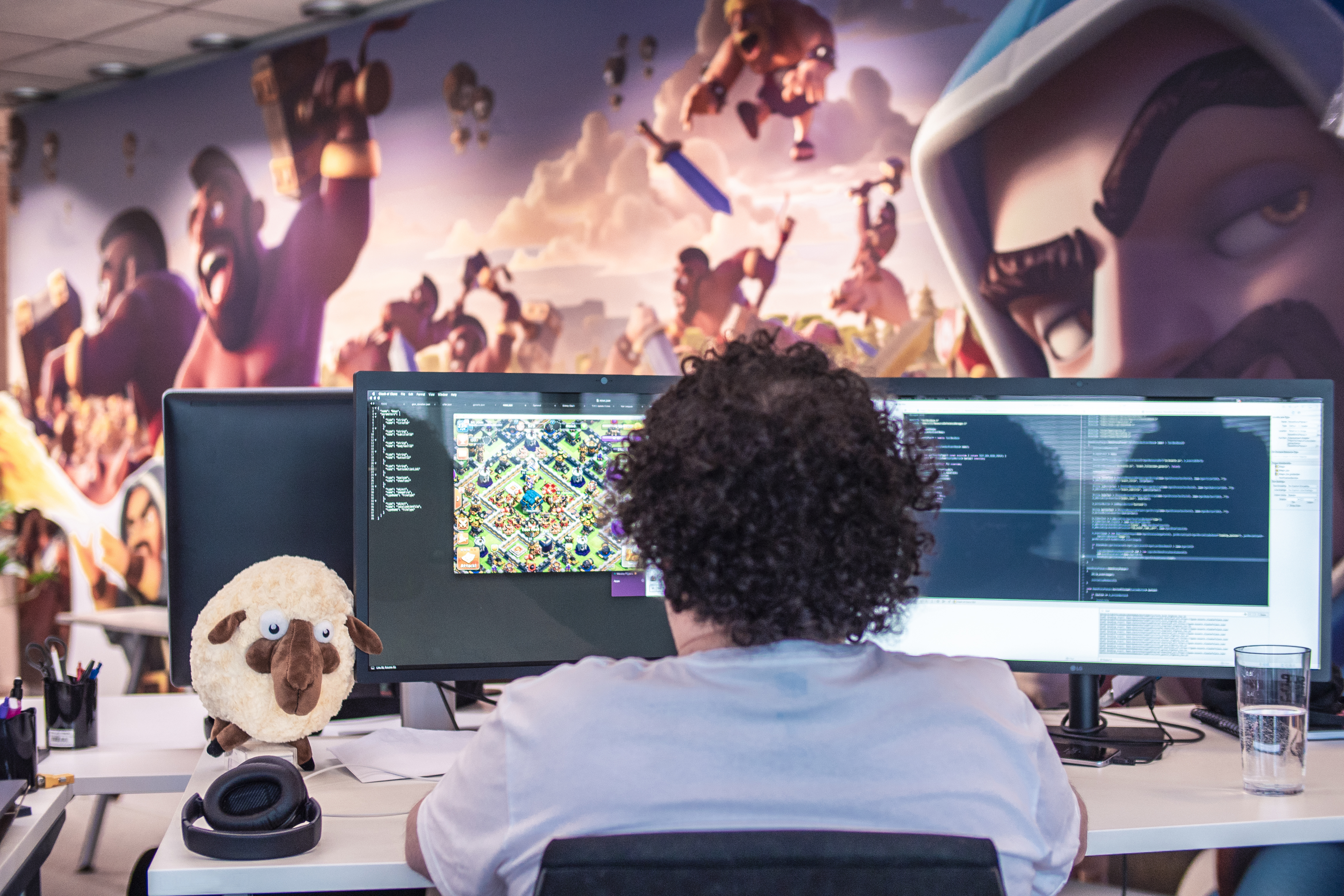 A developer is seen from behind working at a dual monitor setup in a Supercell office, with game code on one screen and game art on the other. The office features colorful game-themed wall art and a plush sheep toy on the desk.