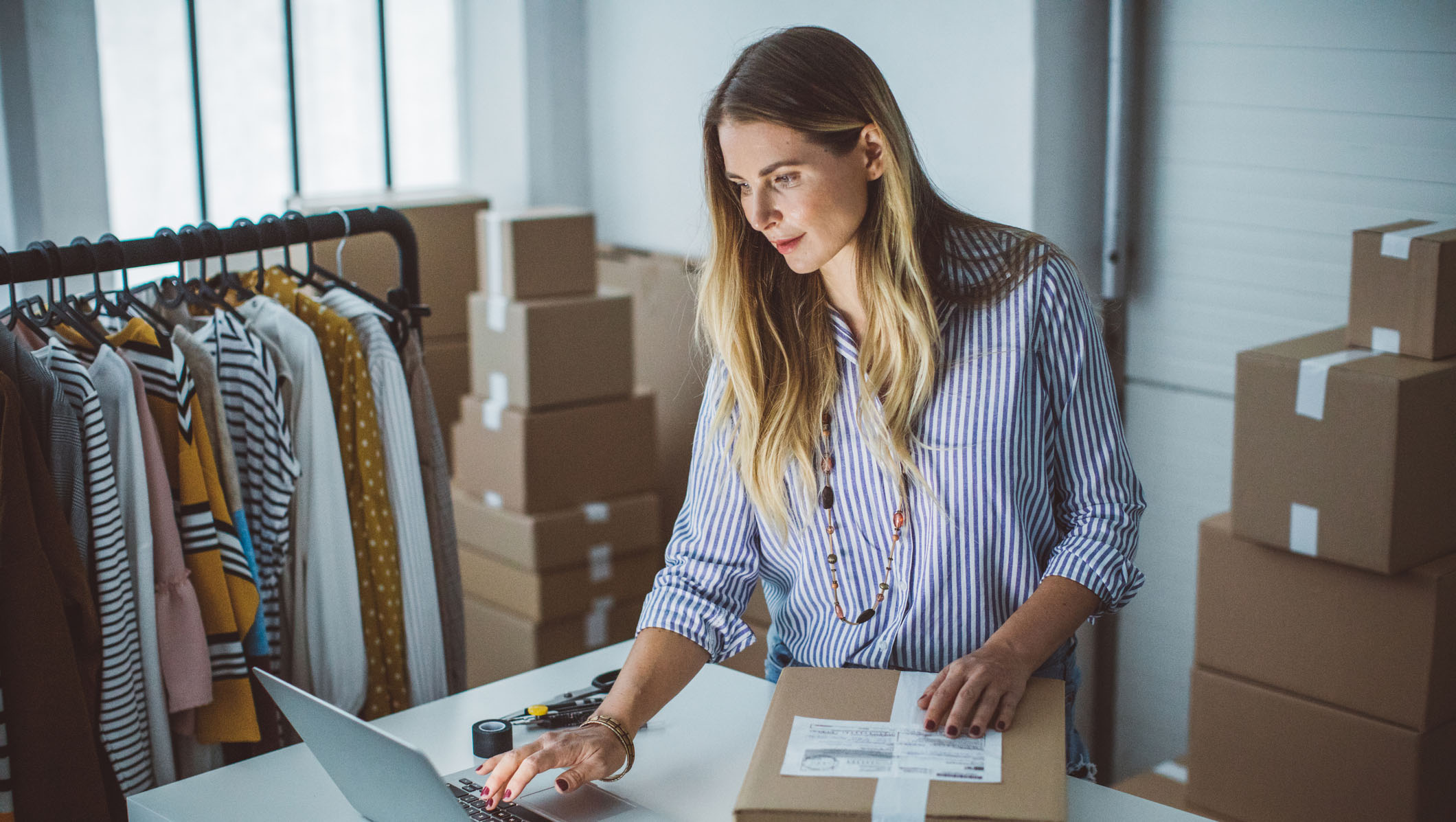 A small business owner prepares and labels a package for delivery while working at a desk with a laptop, surrounded by boxes and clothing products on a rack in a modern workspace.