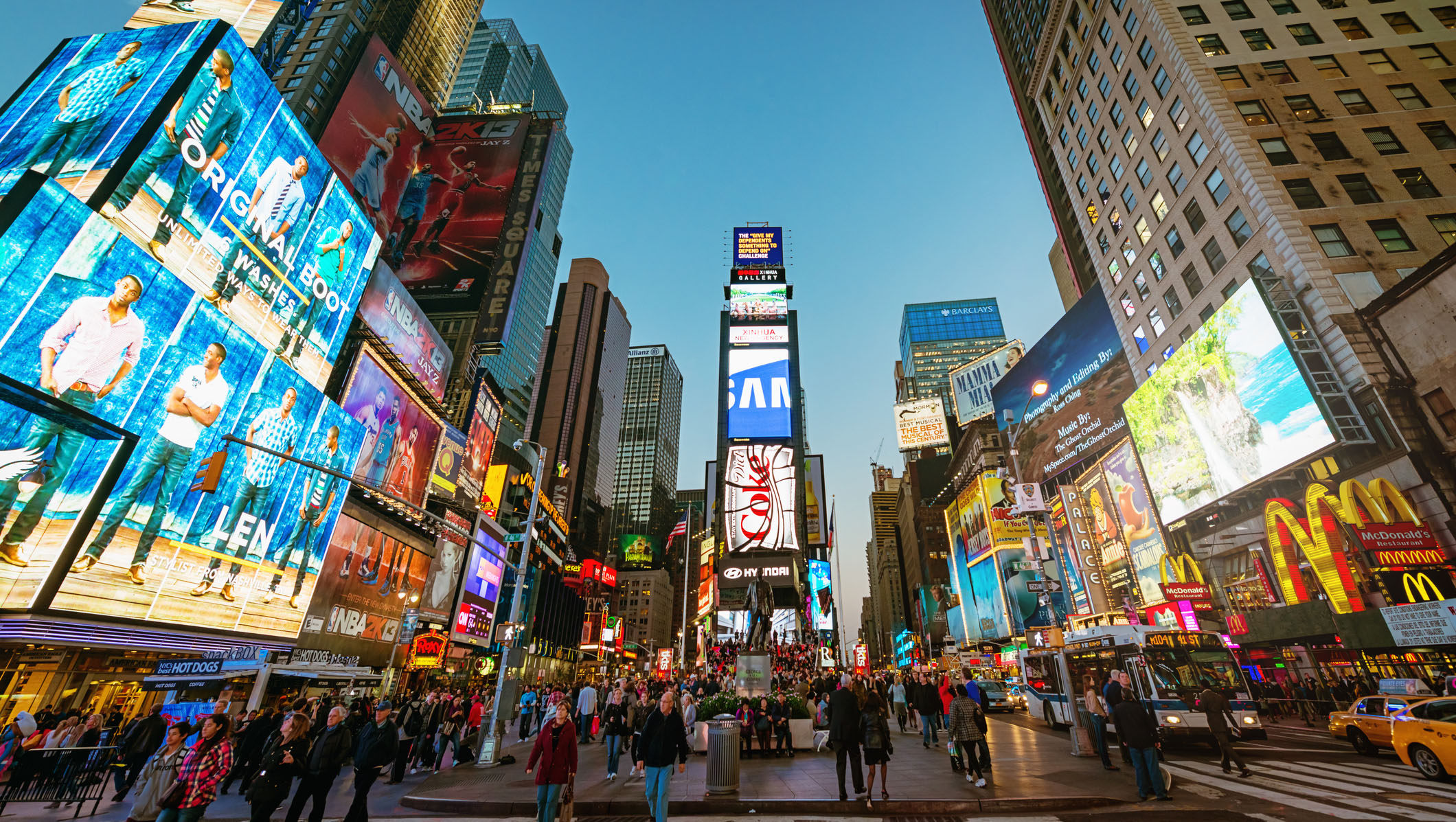 A vibrant scene of Times Square in New York City at dusk, showing crowds of people and buildings adorned with large digital billboards and advertisements.