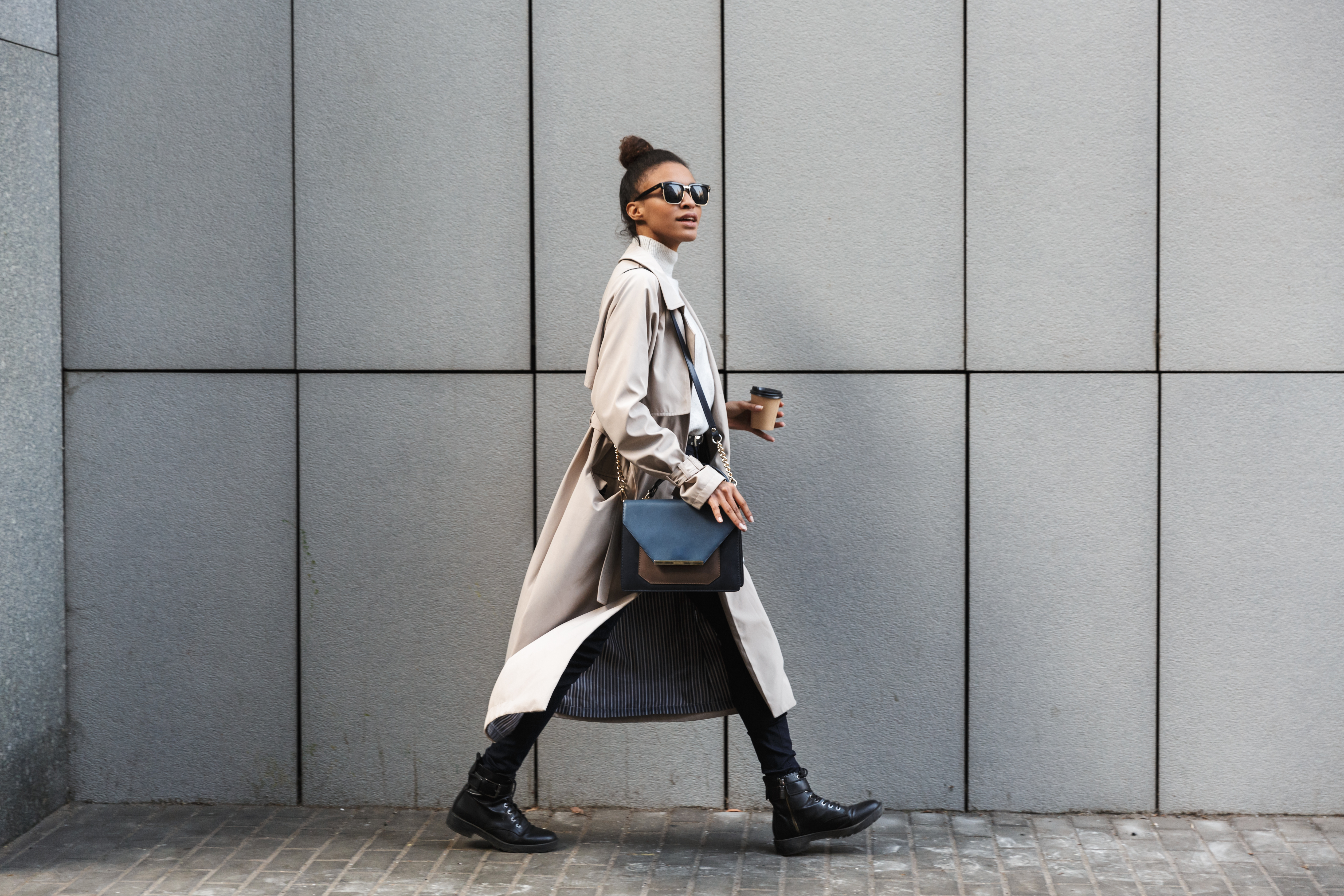 A stylish woman wearing sunglasses and a trench coat walks along a modern city sidewalk while carrying a coffee cup and a handbag.