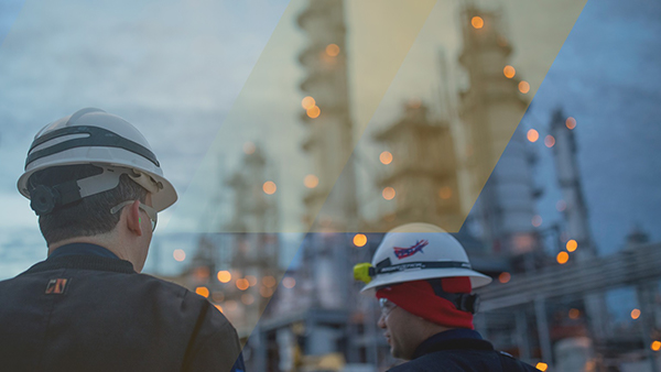 Two engineers wearing safety helmets and protective gear observe equipment at a large industrial facility, with towers and lights in the background.