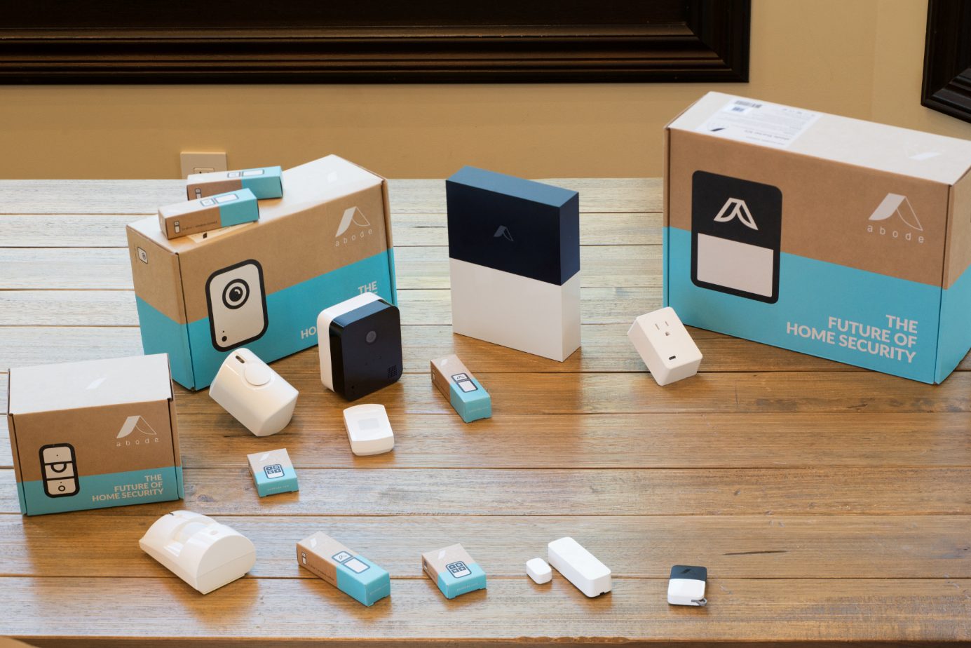A collection of Abode smart home security devices and packaging, including sensors, cameras, outlets, remote controls, and hub units displayed on a wooden table.