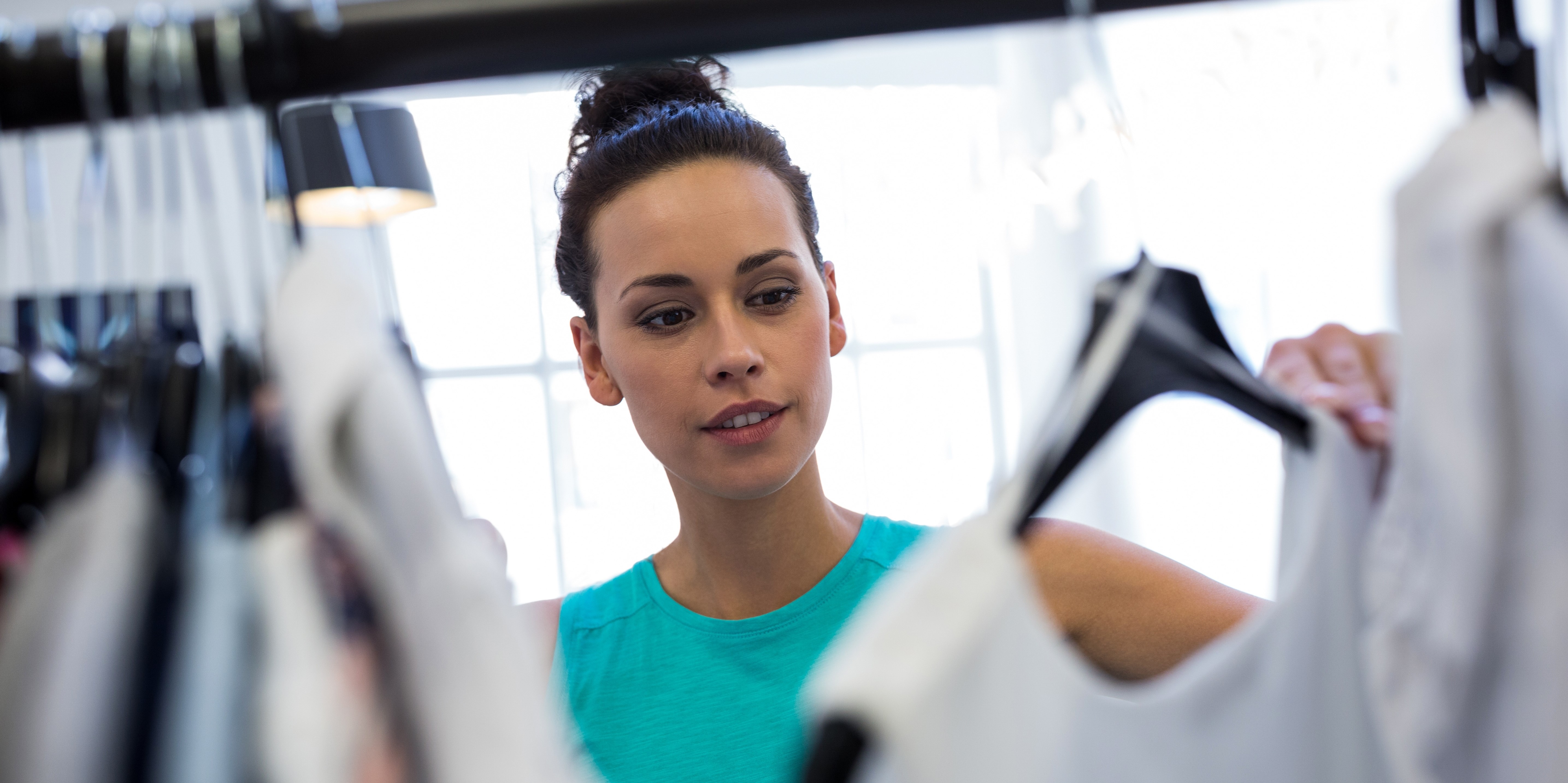 A woman looks through clothes on hangers in a retail store, examining a garment. The image conveys a shopping experience in a bright, modern environment.