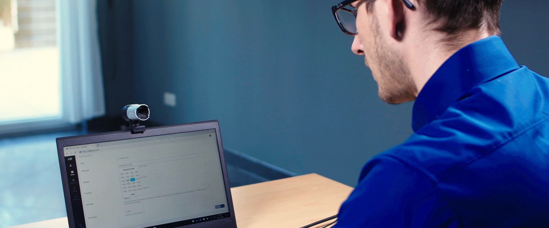 A man wearing glasses and a blue shirt is sitting at a desk in an office, using a laptop with an external webcam attached. He appears to be focused on the screen, possibly participating in a virtual meeting or setting up a video conference.