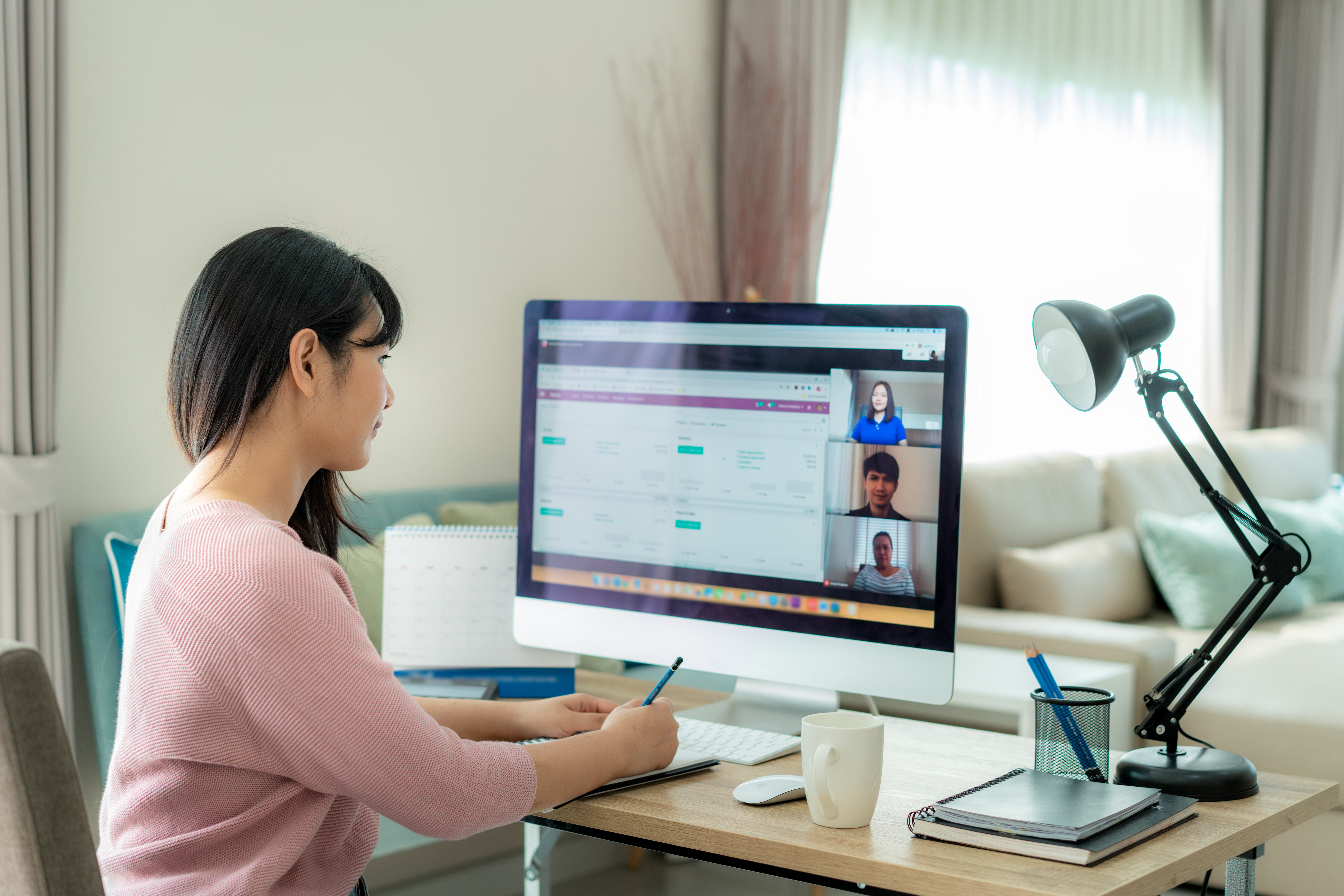 A woman working remotely from her home office, participating in a video conference on a desktop computer. The workspace includes a desk lamp, notebooks, a calendar, and a coffee cup, with a bright living room in the background.
