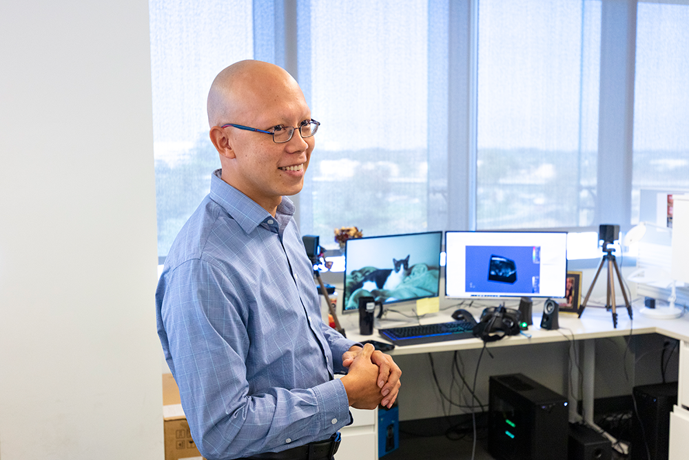 A person standing and smiling in a modern office workspace, with multiple computer monitors and office equipment visible in the background.