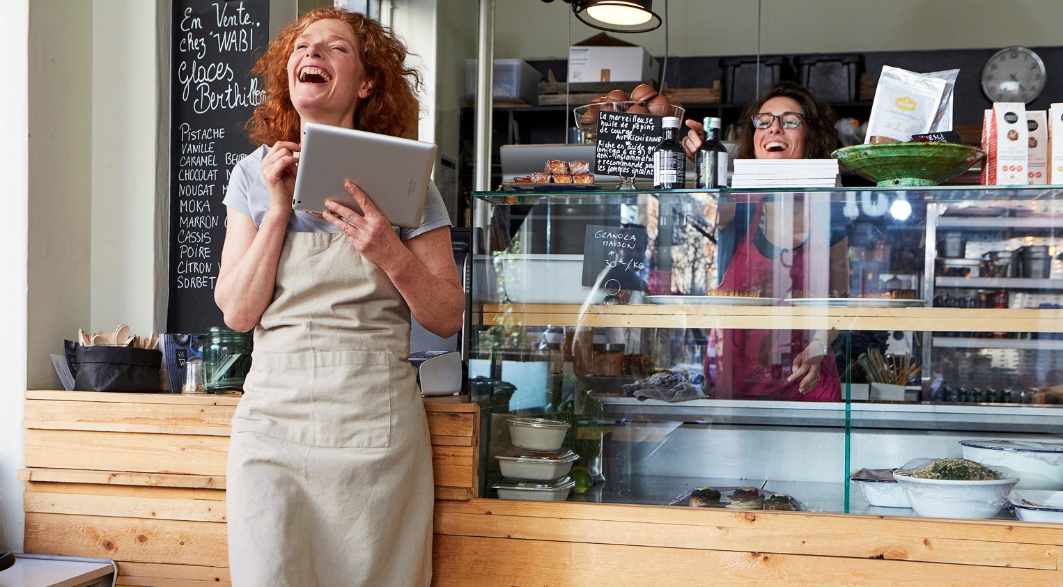 Two women in a café, one wearing an apron and holding a tablet, laughing together behind the counter. The scene highlights small business owners utilizing digital technology in a modern cafe environment.