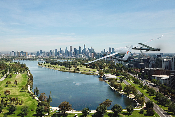 A drone is flying above a large city park with a winding river or lake, modern city buildings, and clear skies in the background.