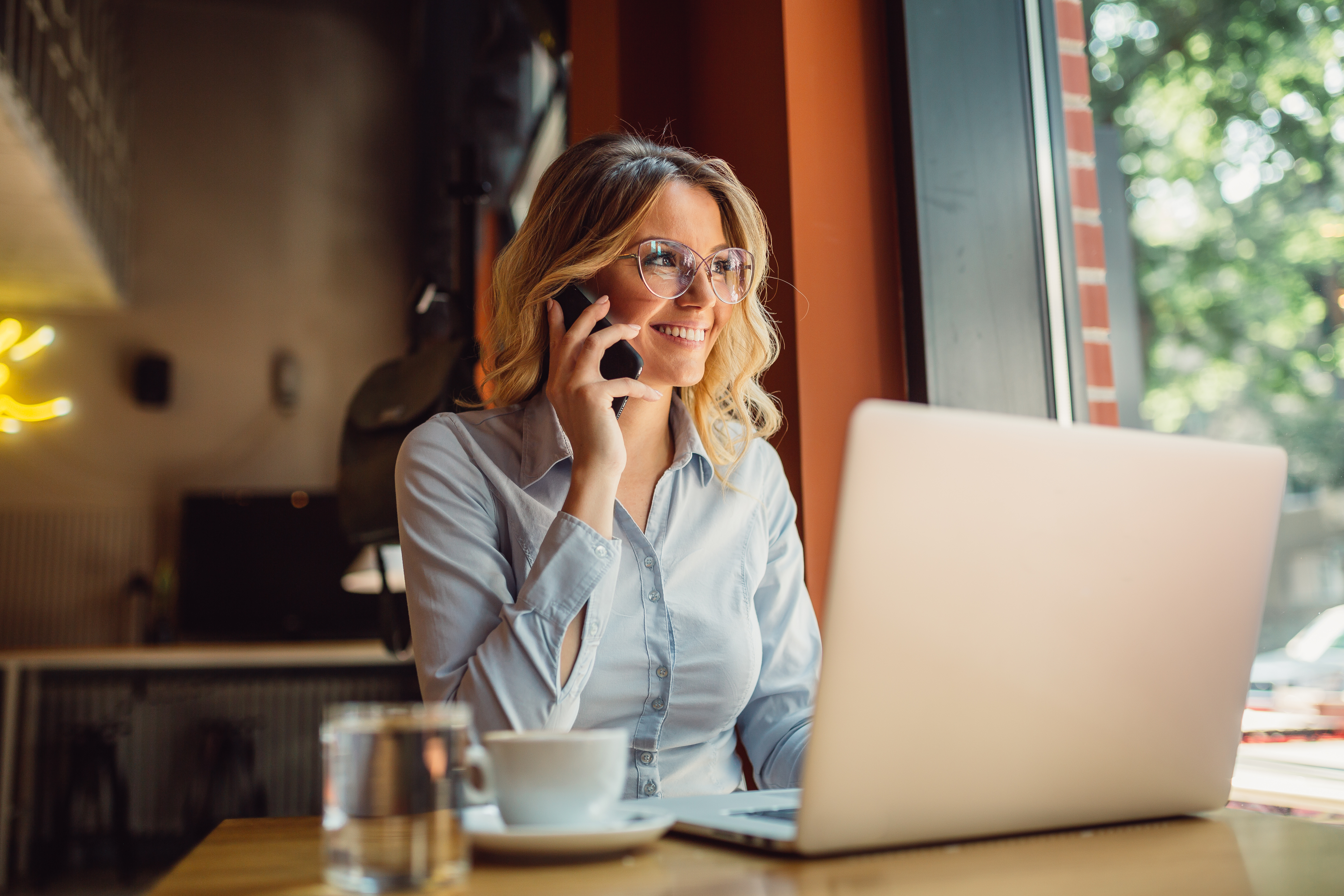 Portrait of young business woman with glasses sitting in cafe in front of her laptop and talking on mobile phone