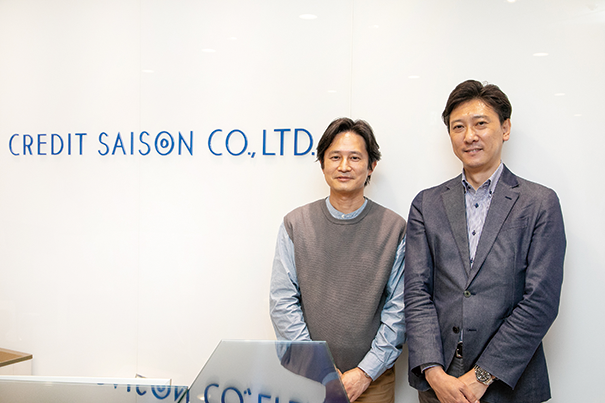 Two representatives stand in front of a Credit Saison Co., Ltd. sign in an office setting.