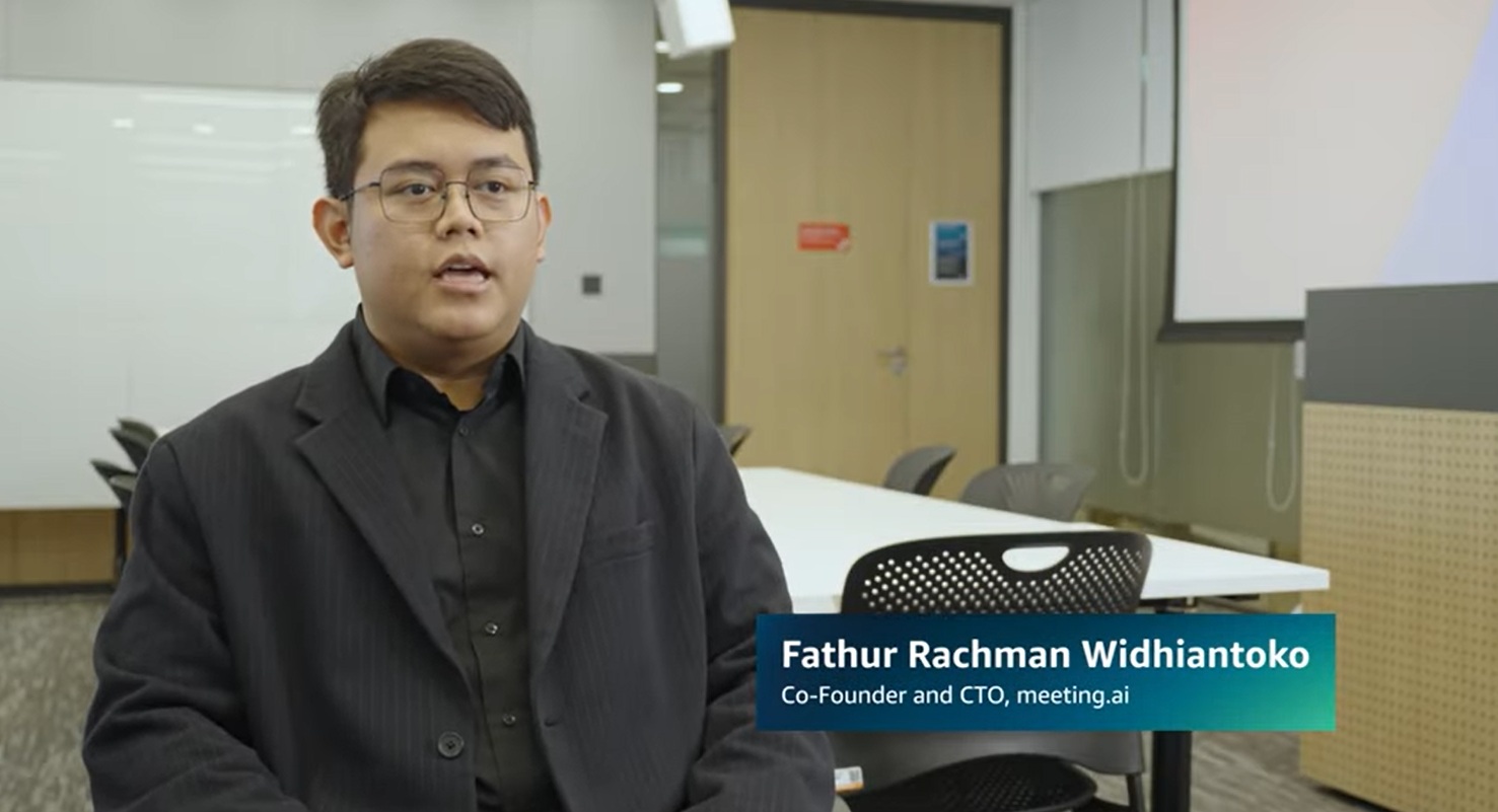 A person wearing glasses and a dark suit is seated for an interview in a modern meeting room. The caption identifies him as Fathur Rachman Widhiantoko, Co-Founder and CTO of meeting.ai. The setting includes tables, chairs, a whiteboard, and a projection screen.