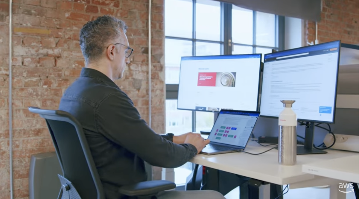 A man working at a desk with a laptop and two large monitors in a modern office with exposed brick walls.