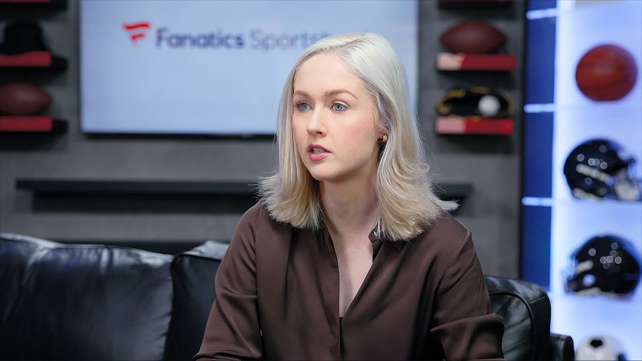 A woman sits on a black sofa in a studio set with sports memorabilia and a sign reading 'Fanatics Sports' in the background. This is a YouTube thumbnail for a sports interview.
