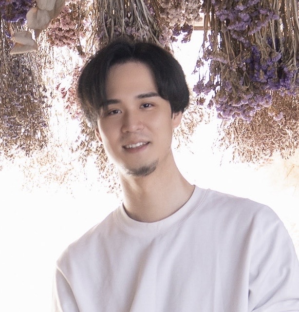 A man in a white shirt standing in front of a background of dried flowers hanging from above.
