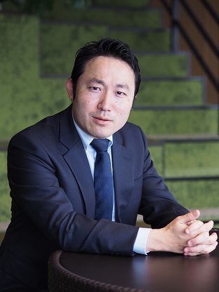 A businessman in a dark suit sits at a round table with his hands folded, with green stairs visible in the background.