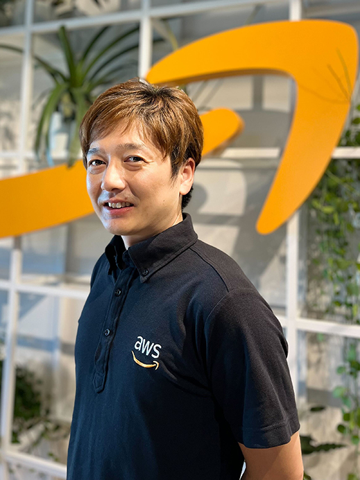 A portrait of an AWS employee standing in an office, wearing an AWS-branded shirt, with the AWS logo visible in the background.