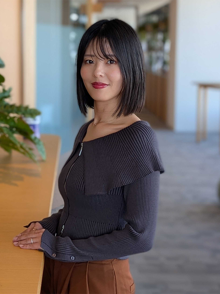 A woman with straight black hair, dressed in a ribbed dark top and brown pants, is standing by a counter in a modern office environment. The setting is bright and professional, with plants and open space visible in the background.