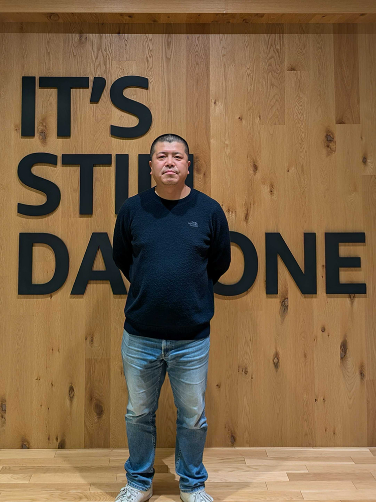 A man stands in front of a wooden wall with the phrase 'IT'S STILL DAY ONE' in large black letters.