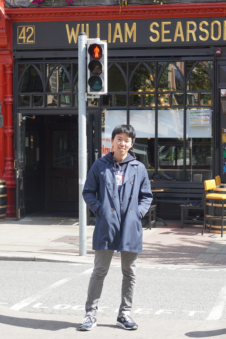 A man wearing a blue coat stands at a crosswalk in front of William Searsons pub, with a red pedestrian traffic light overhead.