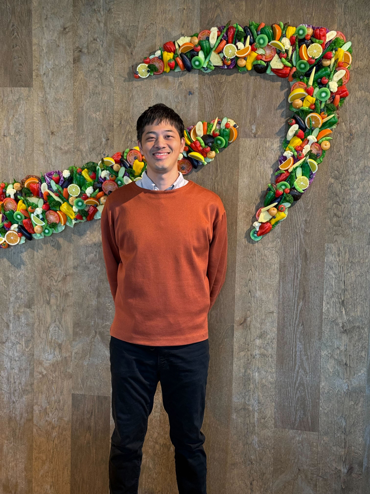 A man wearing an orange sweater is standing and smiling in front of a wall featuring the Amazon logo made out of assorted fruits and vegetables.