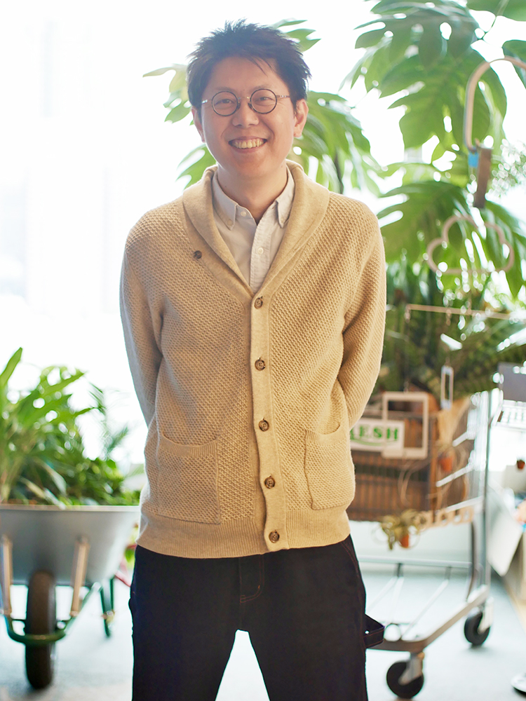 A smiling person wearing a beige cardigan and glasses stands in front of a lush background with various green plants.