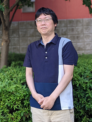 A man wearing glasses and a casual, color-block short-sleeve shirt stands outdoors in front of greenery and a brick wall.