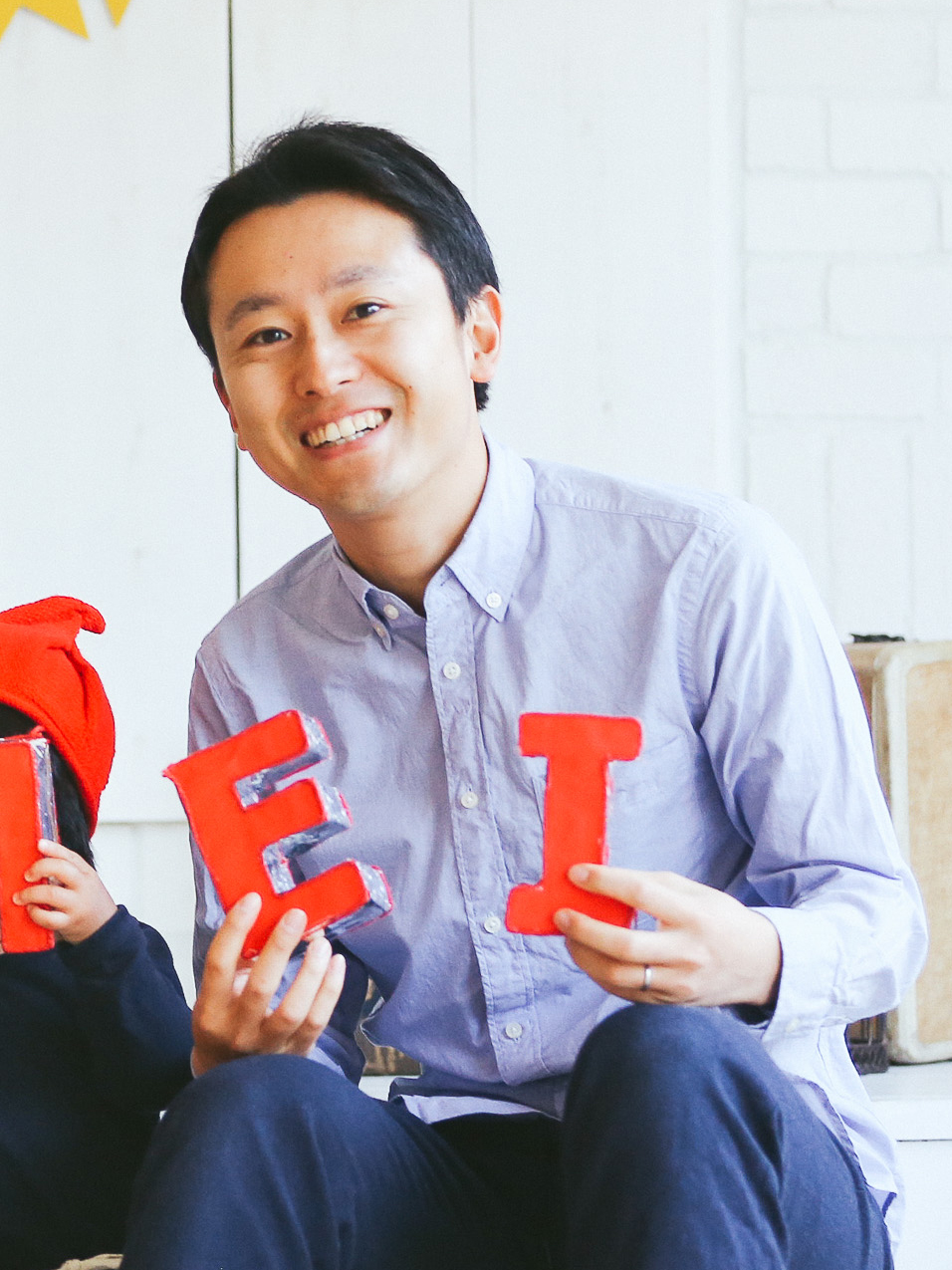 A smiling man in a light blue shirt is holding large red letters E and I. He is seated indoors next to another person, partially visible, who is also holding a red letter.