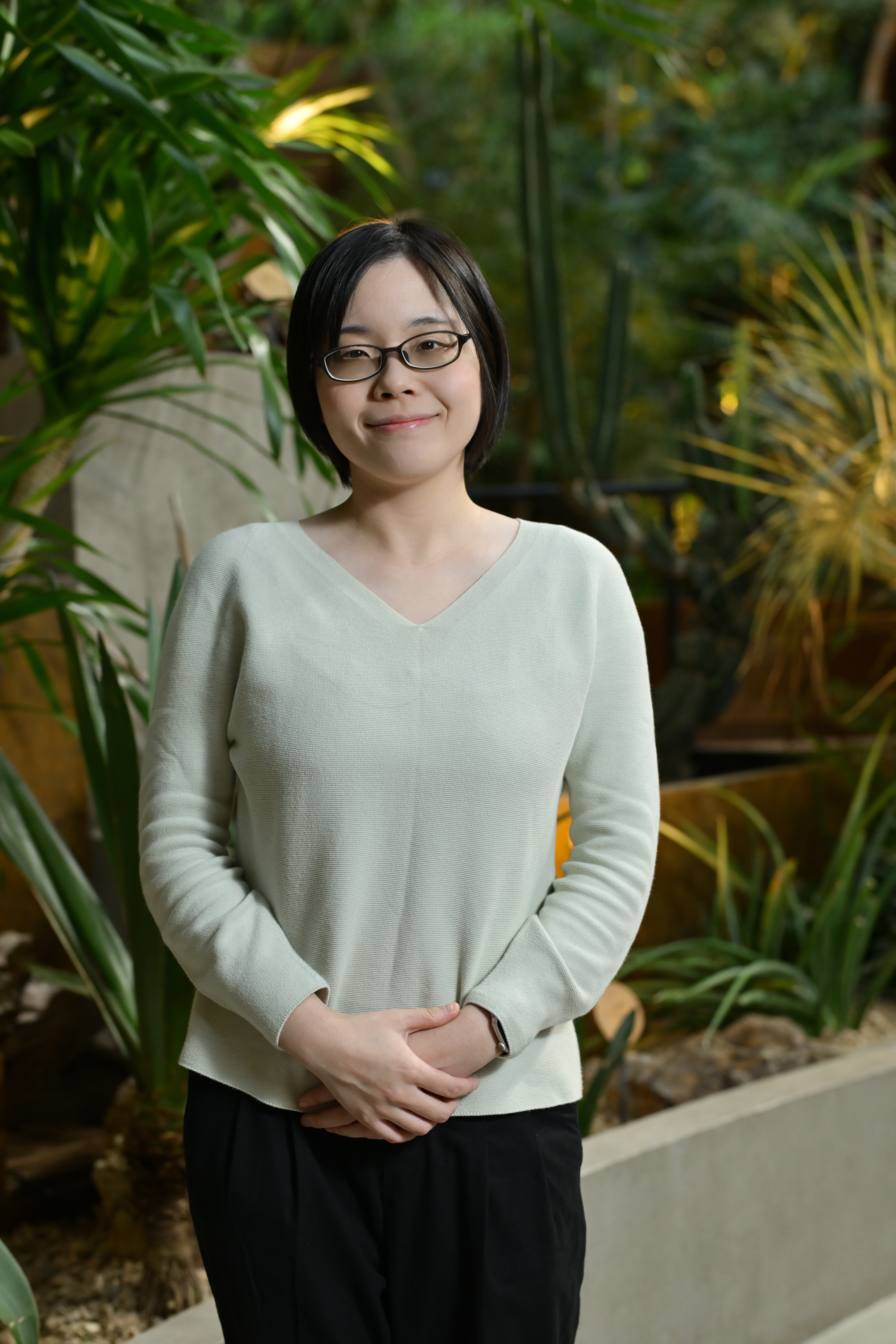 A person wearing a light green sweater and black pants standing in a lush indoor garden with various green plants.