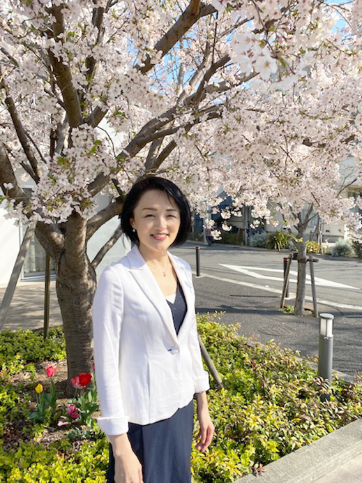 A woman standing outdoors under a blooming cherry blossom tree on a sunny spring day.